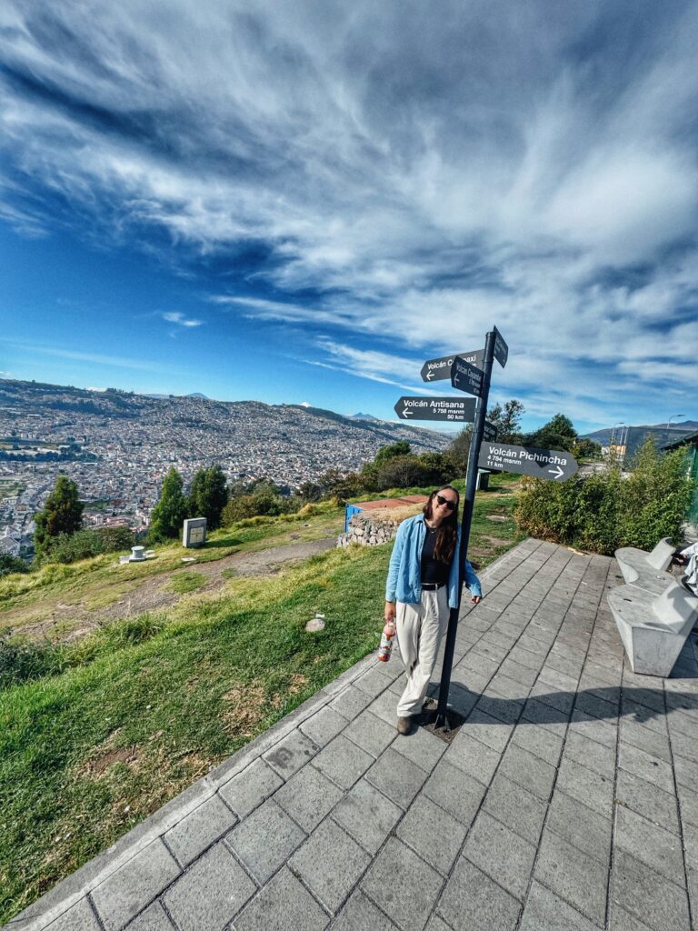 A traveler stands beside a wooden sign in Quito, Ecuador, pointing toward several nearby volcanoes, including Cotopaxi and Pichincha. The Andes Mountains stretch out in the background under a partly cloudy sky.